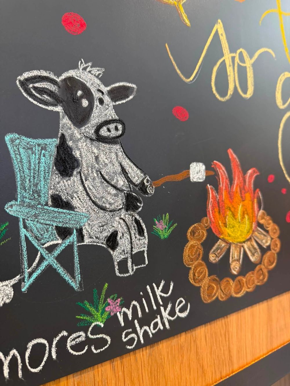 Chalkboard art detail: a spotted cow in a teal camp chair roasting a marshmallow over a campfire, lettered s'mores milkshake