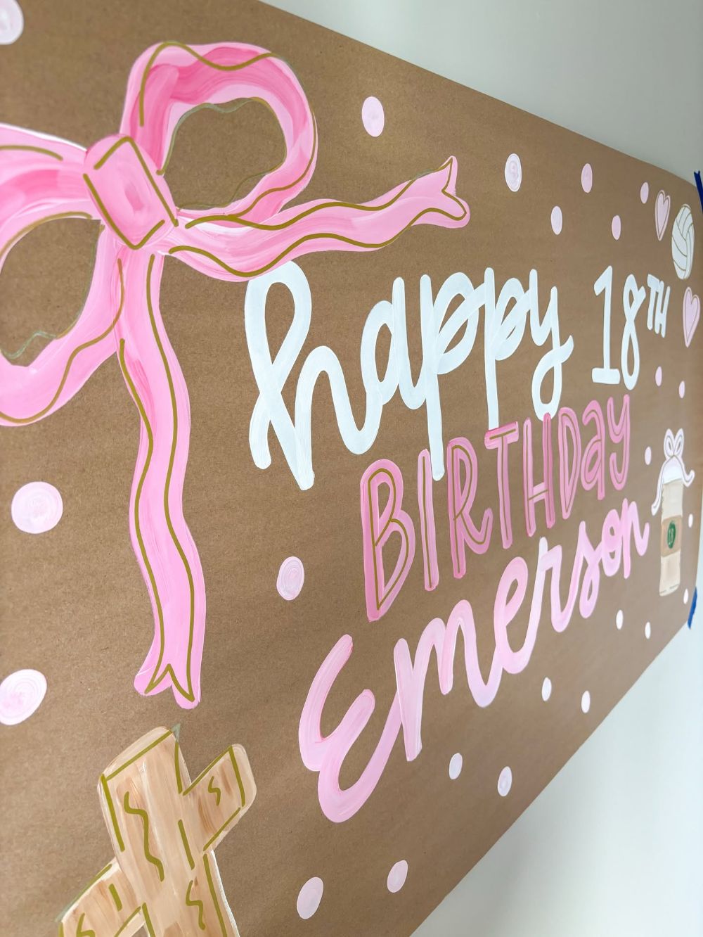 Hand painted 18th birthday banner on kraft paper with a large pink bow, happy 18th birthday Emerson in white and pink lettering, a volleyball, a coffee cup and a cross
