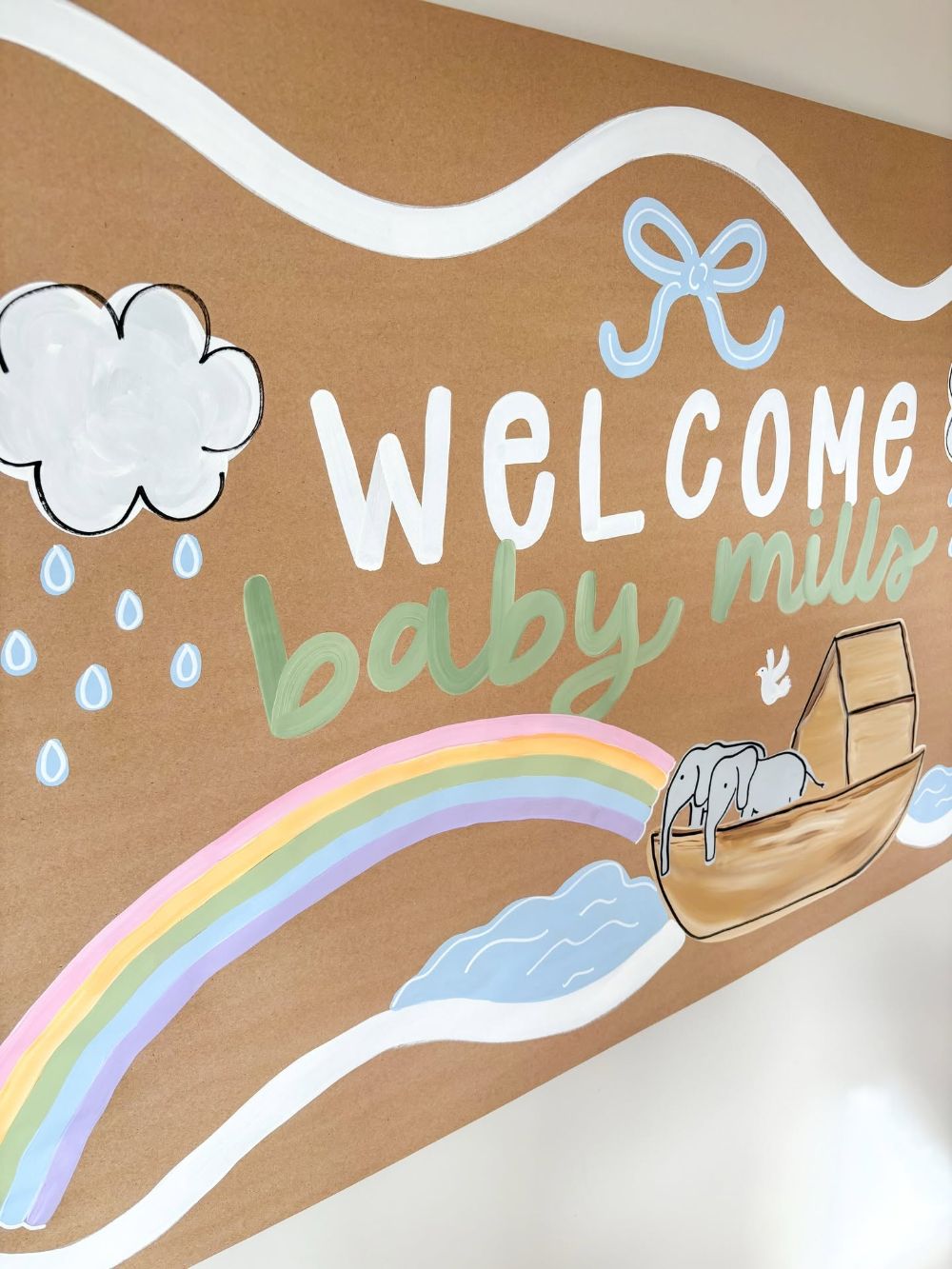 Hand painted baby shower banner with a rainbow, a rain cloud, Noah's ark with two elephants and a dove, lettered welcome baby Mills in white and sage