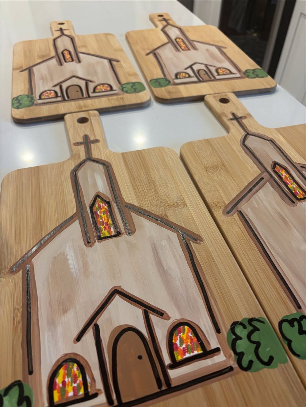 Four bamboo cutting boards hand painted with a white chapel, a cross on the steeple, stained glass windows and green shrubs