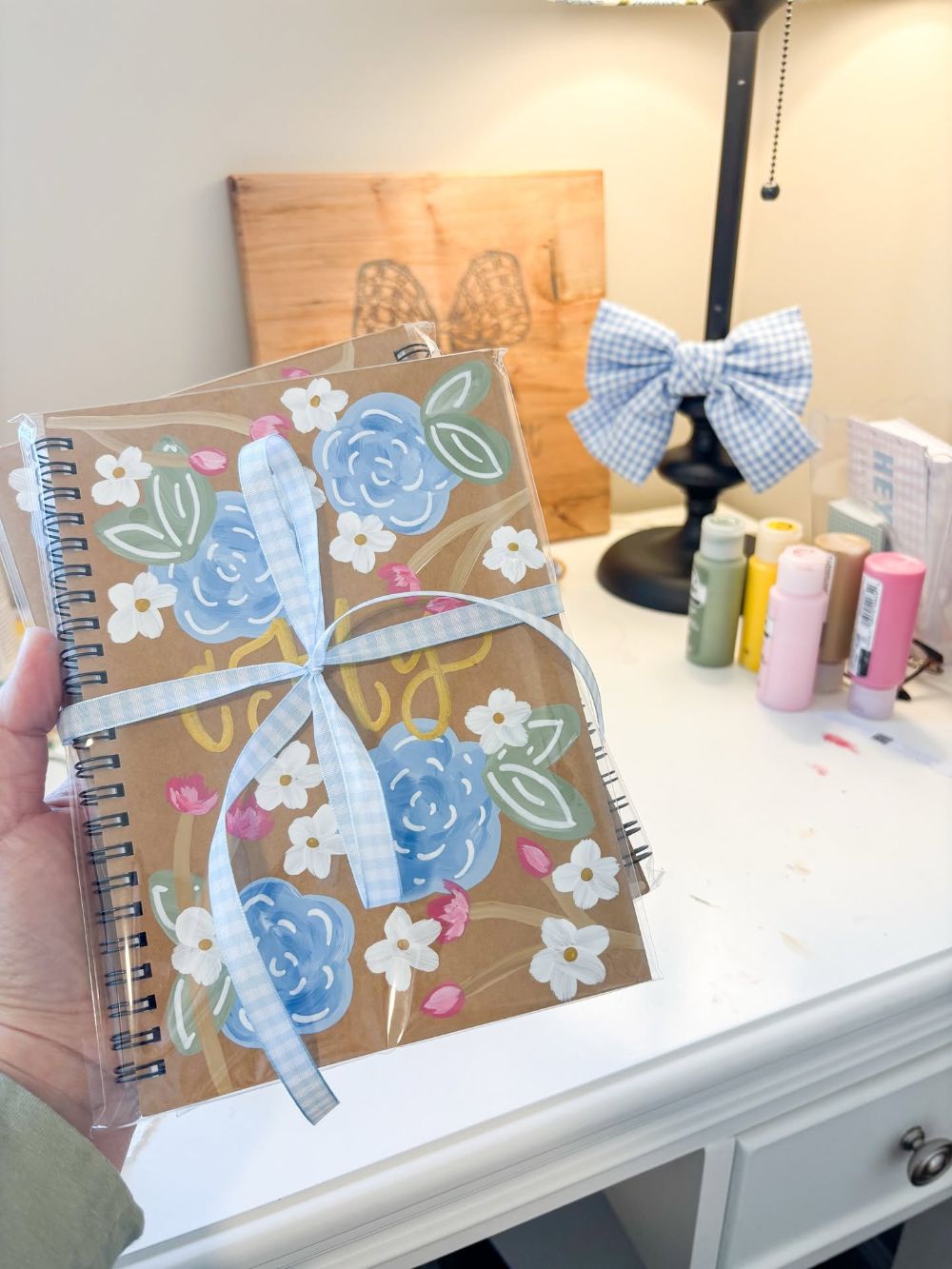 Hand painted kraft notebooks with blue roses, white blossoms and gold lettering, tied with blue gingham ribbon