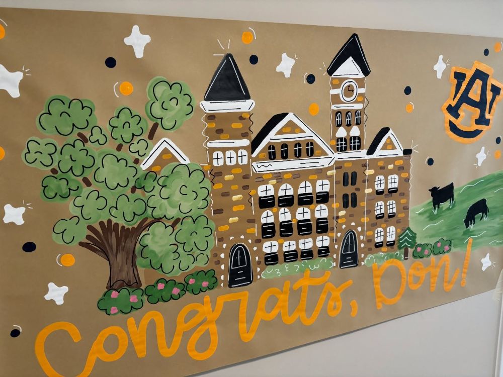 Hand painted graduation banner on kraft paper showing Samford Hall at Auburn University, an oak tree, cows on a hill and the AU logo, lettered congrats, Don in orange