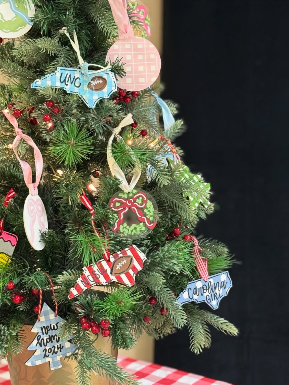 Hand painted North Carolina ornaments on a Christmas tree lettered UNC, NCSU and Carolina girl, with gingham, bow and wreath ornaments and a new home 2024 tree