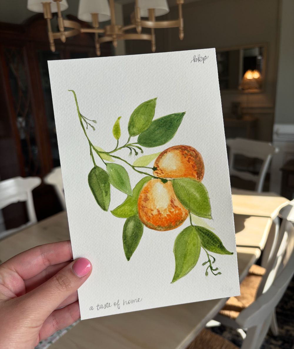 Watercolor painting of two oranges on a leafy branch, hand lettered a taste of home